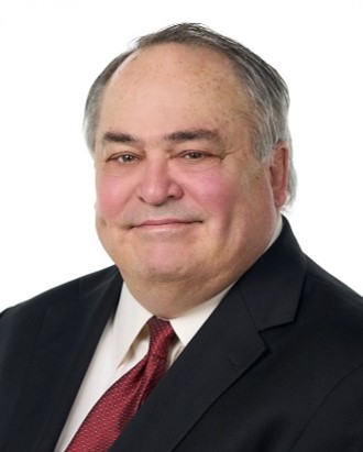 Smirking man with dark eyebrows and balding head with gray hear. He is heavyset and wearing a good jacket with a red tie