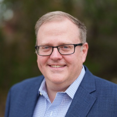 Middle aged man with thinning blonde hair. He smiles at the camera, wearing black square rimmed glasses and a blue suit with a ligh blue checkered shirt