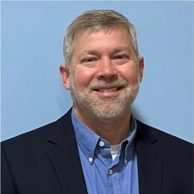 Man with gray hair and short trimmed beard smiles at the camera. He wears a dark blue jacket and lighter blue shirt.