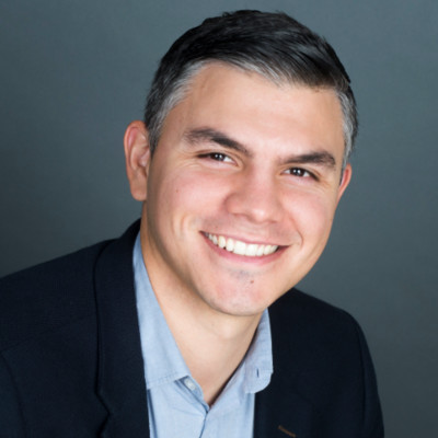 A young man with dark hair smiles at the camera. He has large dark eyebrows and a large smile.